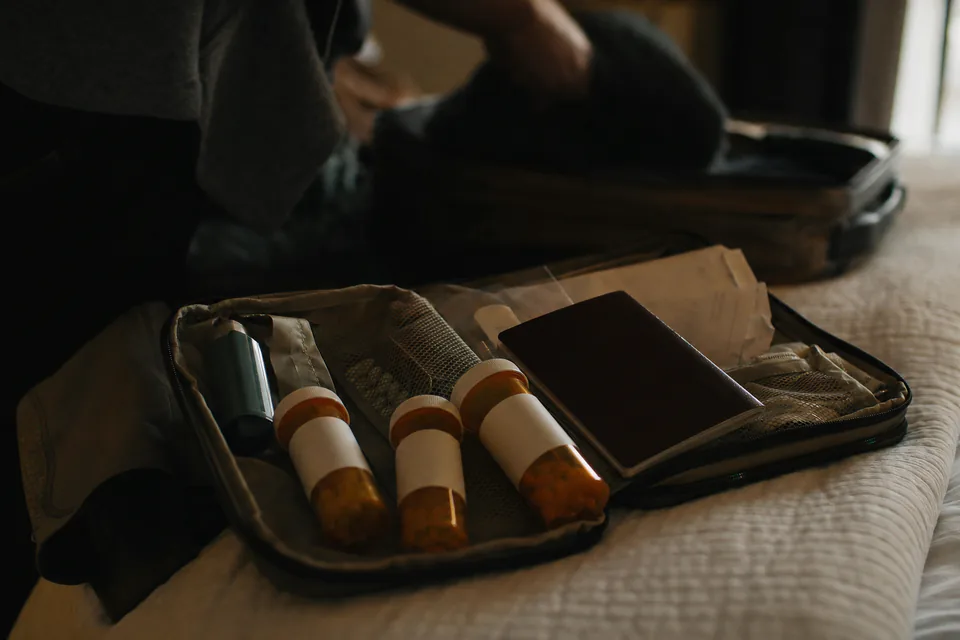 Prescription bottles, an inhaler and a folded doctor letter in an open toiletry bag on a hotel bed
