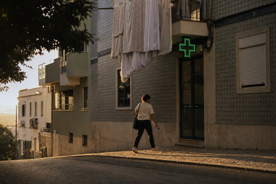 A woman walking along a tiled residential street in Lisbon toward a pharmacy door, its green cross lit above the entrance