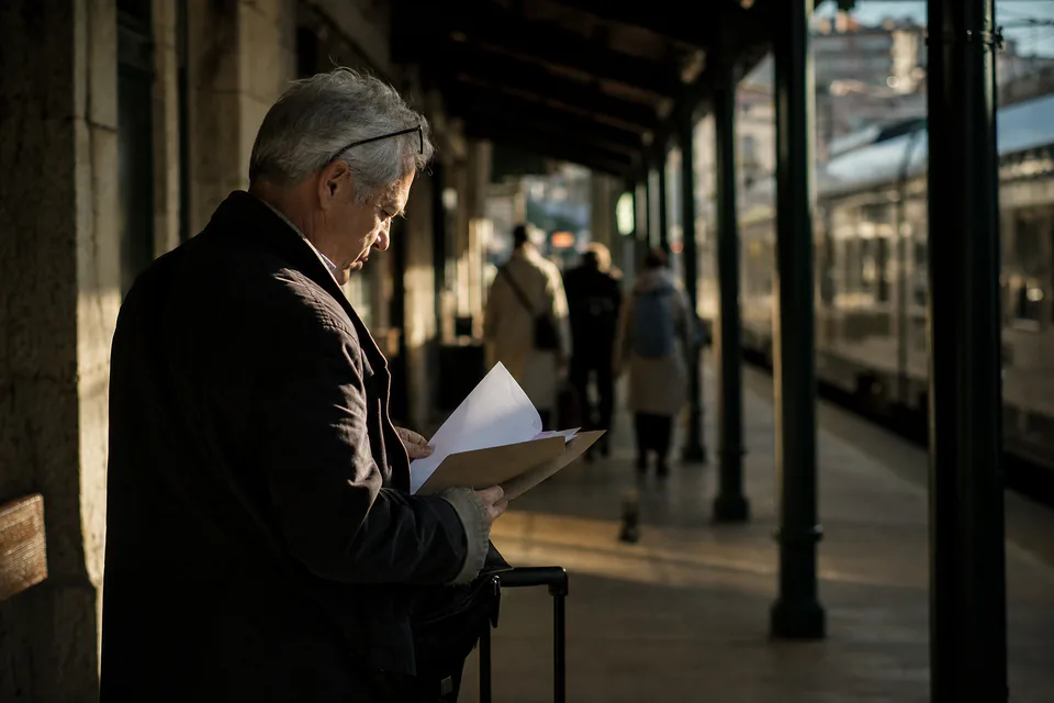 A silver-haired traveller on a railway platform checking a folder of travel documents beside a small suitcase